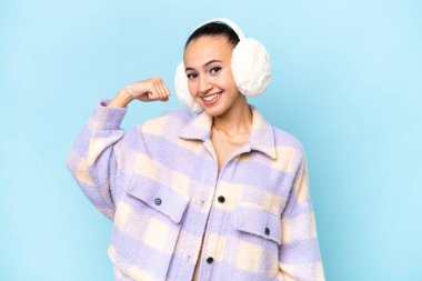 Mavi arka planda izole edilmiş kışlık kuklalar giyen genç bir Arap kadın güçlü bir jest yapıyor.