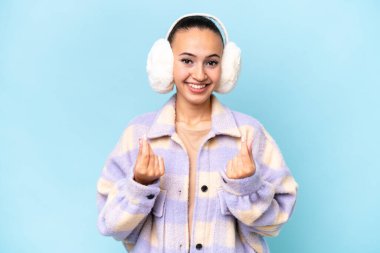 Mavi arka planda izole edilmiş kışlık kuklalar giyen genç bir Arap kadın para hareketi yapıyor.