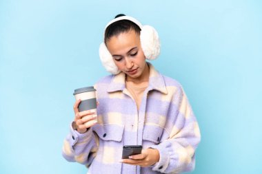 Mavi arka planda izole edilmiş kışlık kuklalar takan genç bir Arap kadın elinde kahve ve bir cep telefonu tutuyor.