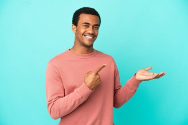 İzole edilmiş mavi arka planda oturan Afro-Amerikalı yakışıklı bir adam bir ilan vermek için avucunda hayali bir şey tutuyor.