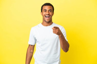 Sarı arka plandaki Afro-Amerikalı yakışıklı adam şaşırmış ve ön tarafı işaret ediyor.