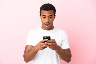 İzole edilmiş pembe arka planda Afro-Amerikalı yakışıklı bir adam kameraya bakarken şaşırmış bir ifadeyle cep telefonuna bakıyor.
