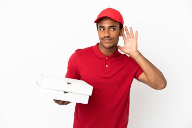 Afro-Amerikalı pizza teslimatçısı izole edilmiş beyaz duvardan pizza kutularını alıyor. Kulağına elini sürerek bir şey dinliyor.