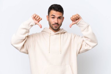 Beyaz arka planda izole edilmiş genç beyaz bir adam baş parmağını iki eliyle aşağı gösteriyor.