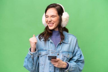 İzole edilmiş arka planda kışlık manşet takan genç beyaz kadın, zafer pozisyonunda telefonla konuşuyor.