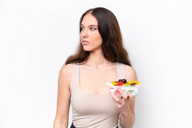 Beyaz arka planda bir kase meyve tutan beyaz bir kadın yan tarafa bakıyor.