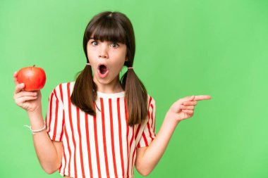 Küçük beyaz kız izole bir arka planda elma tutuyor. Şaşırmış ve işaret ediyor.