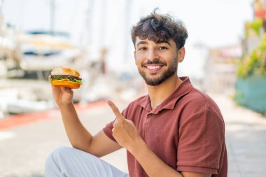 Genç Arap yakışıklı bir adam açık havada bir burger tutuyor ve ona doğrultuyor.