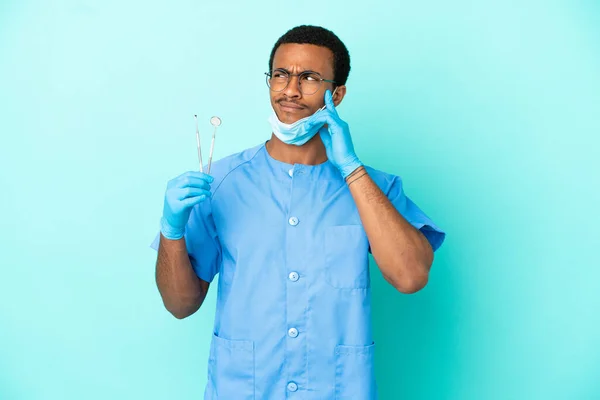 African American dentist holding tools over isolated blue background having doubts and thinking