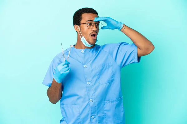 African American dentist holding tools over isolated blue background doing surprise gesture while looking to the side