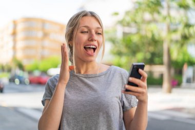 Dışarıdaki genç sarışın kadın kameraya bakarken şaşırmış bir ifadeyle cep telefonunu kullanıyor.