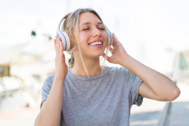 Genç sarışın kadın açık havada müzik dinliyor ve şarkı söylüyor.