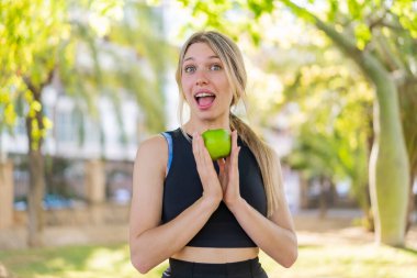 Açık havada genç sarışın bir kadın elinde elmayla şaşkın bir ifadeyle.