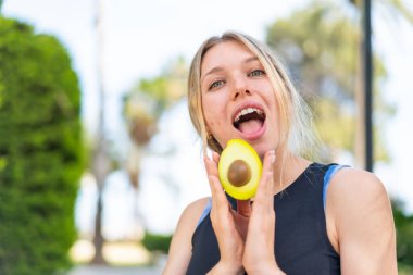 Açık havada genç sarışın bir kadın elinde bir avokado tutuyor. Şaşırmış bir ifadeyle.