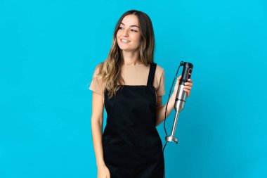 Mavi arka planda el blenderi kullanan Romen kadın yana bakıyor ve gülümsüyor