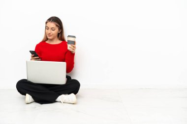 Dizüstü bilgisayarı olan, elinde kahve, elinde cep telefonu olan genç, beyaz bir kadın.