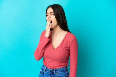 Genç beyaz kadın mavi arka planda izole edilmiş esniyor ve geniş bir ağzı eliyle kapatıyor.