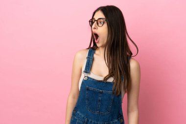 Pembe arka planda izole edilmiş genç beyaz kadın yan tarafa bakarken sürpriz jest yapıyor.