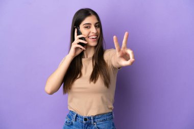 Mor arka planda izole edilmiş cep telefonu kullanan beyaz bir kadın gülümsüyor ve zafer işareti gösteriyor.