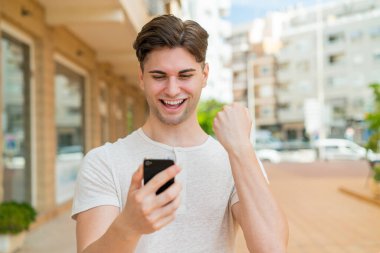 Genç yakışıklı adam cep telefonu kullanıyor ve zafer jesti yapıyor.