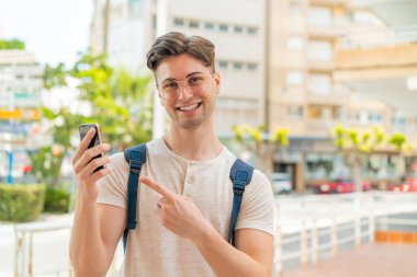 Genç ve yakışıklı bir adam açık havada cep telefonu kullanıyor ve onu işaret ediyor.