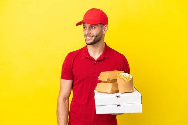 Pizza teslimatçısı, izole edilmiş arka planda pizza kutularını ve burgerleri alıp yan tarafa bakıyor ve gülümsüyor.
