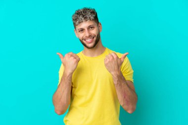 Mavi arka planda soyutlanmış yakışıklı beyaz bir adam. Baş parmakları havada ve gülümsüyor.
