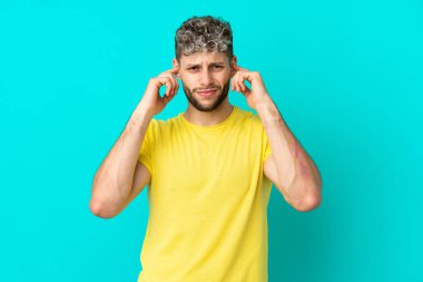 Mavi arka planda izole edilmiş yakışıklı beyaz adam hayal kırıklığına uğramış ve kulaklarını kapatmış.