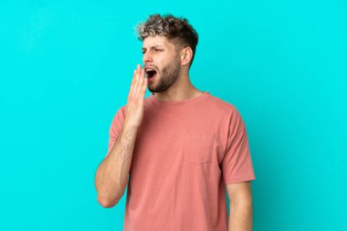 Mavi arka planda izole edilmiş yakışıklı beyaz adam esniyor ve ağzını eliyle açık tutuyor.