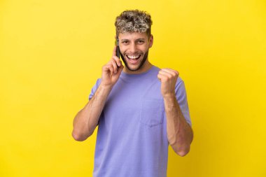 Sarı arka planda izole edilmiş cep telefonu kullanan genç beyaz adam galibiyeti kutluyor.