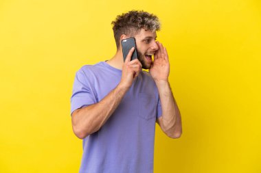 Sarı arka planda izole edilmiş cep telefonu kullanan genç beyaz adam ağzı sonuna kadar açık bağırıyor.
