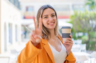 Genç ve güzel bir kadın elinde kahveyle dışarıda gülümsüyor ve zafer işareti gösteriyor.