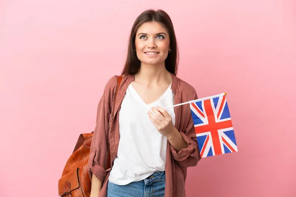 年轻的高加索女人举着一面英国国旗 背景是粉色的 一边高瞻远瞩 一边思考问题 — 图库照片