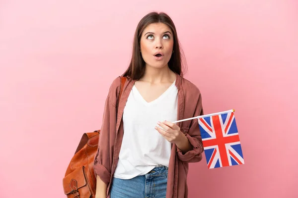 年轻的高加索女人举着一面英国国旗 背景是粉色的 抬起头 表情惊讶 — 图库照片