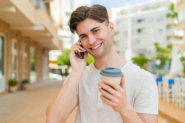 Młody Przystojny Mężczyzna Używa Telefonu Komórkowego Trzyma Kawę Szczęśliwą Ekspresją — Zdjęcie stockowe