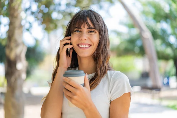 屋外で携帯電話を使用し 幸せな表情のコーヒーを保持する若い女性 — ストック写真