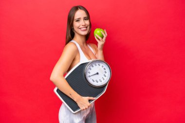 Young beautiful caucasian woman isolated on red background with weighing machine and with an apple