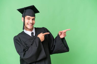 Tecrit edilmiş arka planda genç üniversite mezunu beyaz bir adam parmak göstererek bir ürün sunuyor.