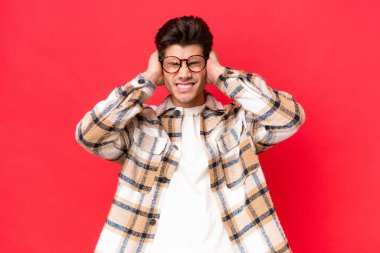 Kızıl arka planda izole edilmiş genç beyaz yakışıklı adam hayal kırıklığına uğramış ve kulaklarını kapatmış.