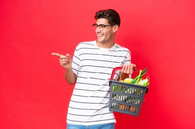 Kafkasyalı genç bir adam elinde yiyecek dolu bir sepet tutuyor. Kırmızı arka planda izole edilmiş. Parmakla göstererek bir ürün sunuyor.