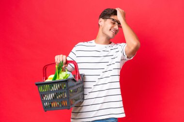 Kafkasyalı genç bir adam elinde kırmızı arka planda izole edilmiş yiyecek dolu bir sepet tutuyor. Bir şey fark etti ve çözümü arıyor.