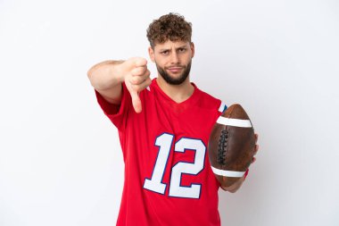 Beyaz arka planda ragbi oynayan beyaz tenli genç bir adam olumsuz bir ifadeyle baş parmağını aşağı gösteriyor.
