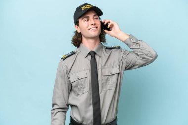 Genç güvenlik görevlisi mavi arka planda izole edilmiş cep telefonuyla sohbet ediyor.
