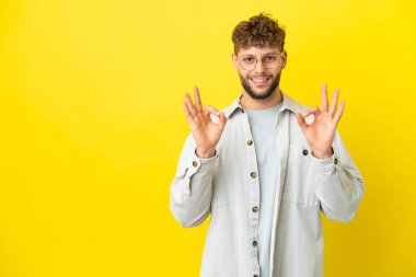 Sarı arka planda izole edilmiş yakışıklı beyaz adam iki eliyle tamam işareti gösteriyor.