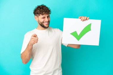 Genç beyaz adam mavi arka planda izole edilmiş. Elinde yeşil işaret simgesi olan bir pankart var. Ve ön tarafı işaret ediyor.
