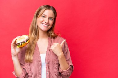 Young caucasian woman holding a burger  isolated on red background pointing to the side to present a product