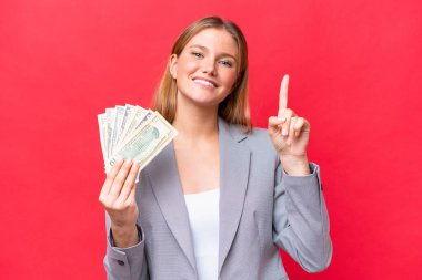 Kafkasyalı genç bir kadın kırmızı arka planda izole edilmiş para tutuyor ve en iyinin işareti olarak parmağını kaldırıyor.