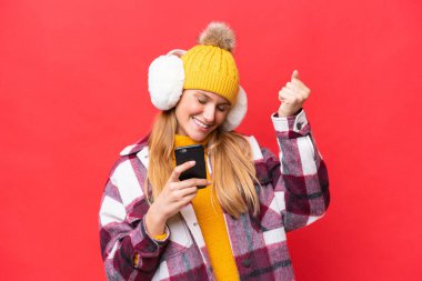 Kırmızı arka planda izole edilmiş kışlık kürkler giyen genç ve güzel bir kadın ve telefon zafer pozisyonunda.