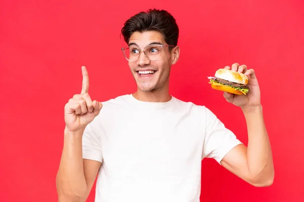 Młody Biały Mężczyzna Trzymając Hamburgera Izolowany Czerwonym Tle Zamiarem Realizacji — Zdjęcie stockowe