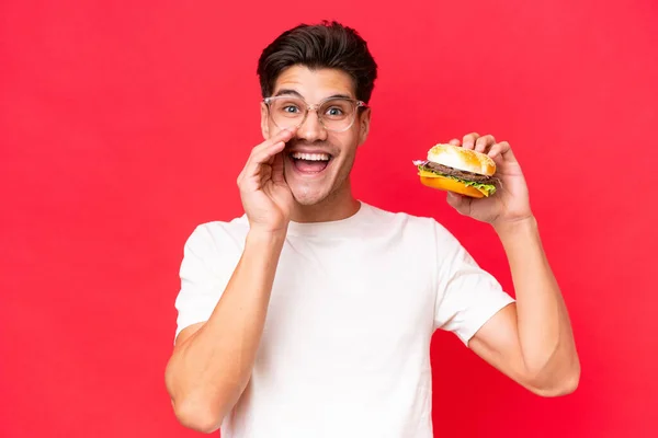 Młody Biały Mężczyzna Trzyma Hamburgera Odizolowany Czerwonym Tle Krzycząc Szeroko — Zdjęcie stockowe